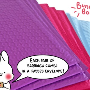 Puede incluir: Sobres acolchados de color morado, rosa y azul con el texto "&iexcl;Cada par de pendientes viene en un sobre acolchado!" y un conejito de dibujos animados que sostiene un sobre peque&ntilde;o con un coraz&oacute;n. El texto "Bunny Boop" est&aacute; escrito con una fuente divertida encima de los sobres.