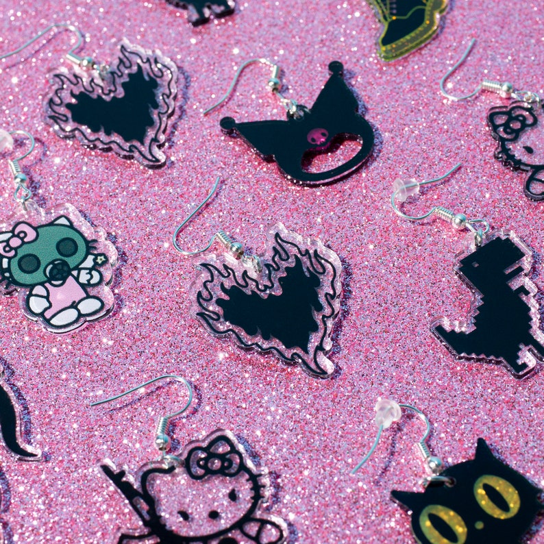 Puede incluir: Una colecci&oacute;n de pendientes de acr&iacute;lico blanco y negro con varios dise&ntilde;os, incluyendo un coraz&oacute;n con llamas, un dinosaurio, un gato y un personaje de Hello Kitty. Los pendientes se muestran sobre un fondo de purpurina rosa.
