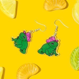 May include: A pair of dangle earrings featuring a green frog wearing a pink cowboy hat. The earrings are made of clear acrylic and have silver hooks.