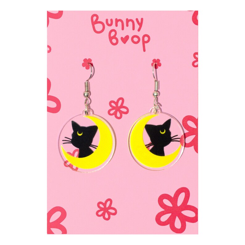 Puede incluir: Un par de pendientes de gato negro y luna creciente. Los pendientes est&aacute;n hechos de acr&iacute;lico transparente con una luna creciente amarilla y una silueta de gato negro. Los pendientes est&aacute;n sobre un fondo rosa con el texto "Bunny Boop" en blanco.