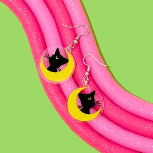 Puede incluir: Un par de pendientes de plata con siluetas de gatos negros sobre medias lunas amarillas. Los pendientes est&aacute;n sobre un fondo rosa y blanco.