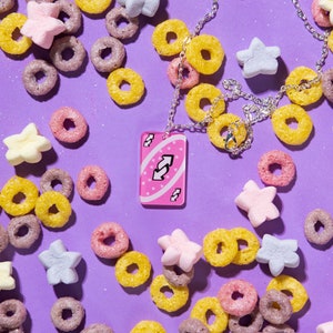 May include: A pink Uno card necklace with a reverse arrow symbol. The necklace is surrounded by cereal and marshmallows on a purple background.