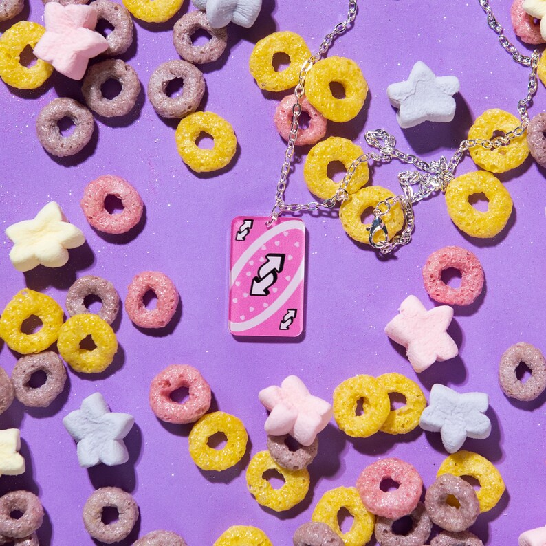 May include: A pink Uno card necklace with a reverse arrow symbol. The necklace is surrounded by cereal and marshmallows on a purple background.