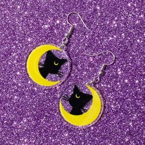 Puede incluir: Un par de pendientes de plata con una silueta de gato negro sobre una luna creciente amarilla. Los pendientes est&aacute;n sobre un fondo de purpurina morado.