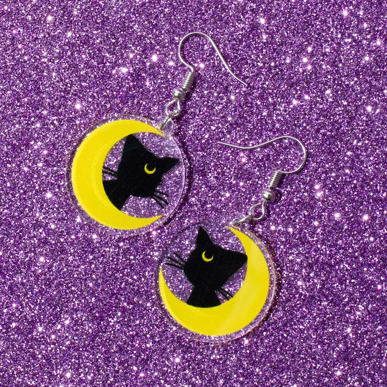 Puede incluir: Un par de pendientes de plata con una silueta de gato negro sobre una luna creciente amarilla. Los pendientes est&aacute;n sobre un fondo de purpurina morado.