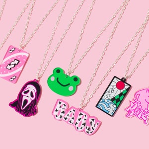 May include: Seven silver chains with different charms hanging from them. The charms are a pink and white card with a swirl design, a pink and black ghost face, a green frog, a pink and black "BABY" charm, a pink and black Japanese-style design, and a pink and black "RARE" charm.