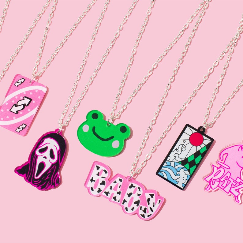May include: Seven silver chains with different charms hanging from them. The charms are a pink and white card with a swirl design, a pink and black ghost face, a green frog, a pink and black "BABY" charm, a pink and black Japanese-style design, and a pink and black "RARE" charm.