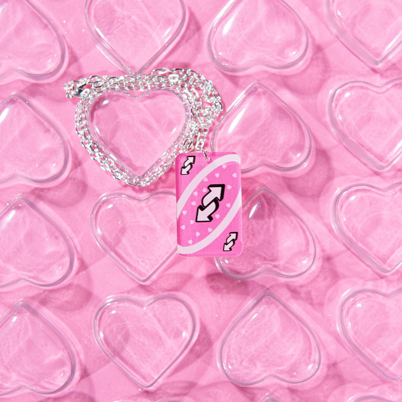 May include: A pink Uno card necklace with a silver chain. The card features the reverse symbol and is surrounded by white polka dots. The necklace is displayed on a pink background with heart-shaped cutouts.