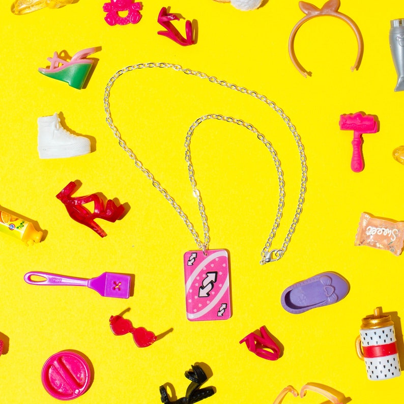 May include: A silver chain necklace with a pink Uno card pendant featuring a double arrow symbol. The necklace is surrounded by various colorful accessories, including shoes, hair accessories, and a spatula.