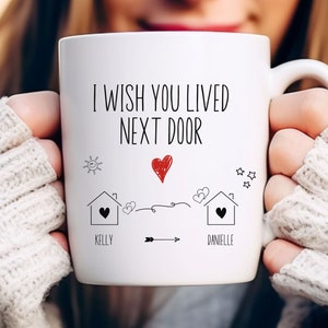 May include: White ceramic mug with a black and red design. The mug says "I wish you lived next door" with a drawing of two houses, a heart, and an arrow. The names "Kelly" and "Danielle" are written below the houses.