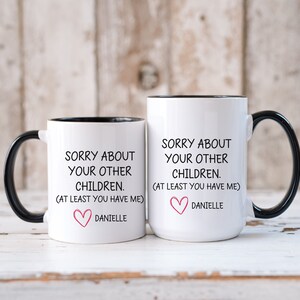 May include: Two white coffee mugs with black handles and black rims. The mugs have a funny message printed on them that says "Sorry about your other children. (At least you have me)" with a pink heart and the name Danielle below.