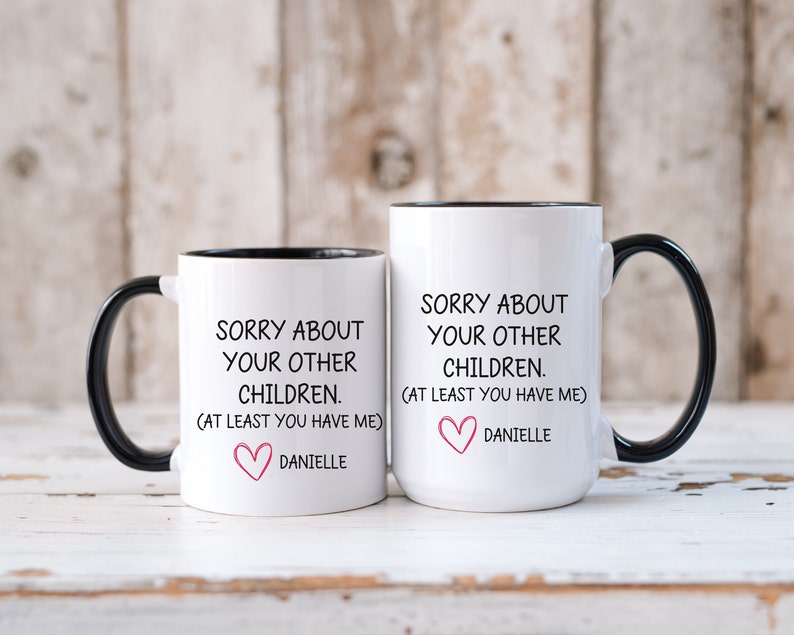 May include: Two white coffee mugs with black handles and black rims. The mugs have a funny message printed on them that says "Sorry about your other children. (At least you have me)" with a pink heart and the name Danielle below.