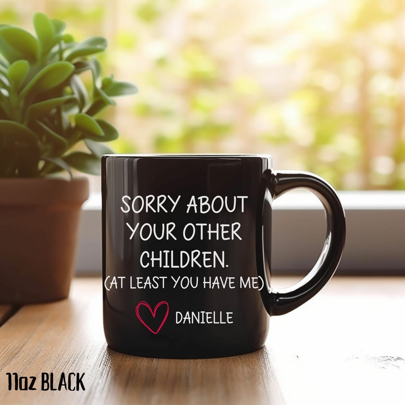 May include: Black ceramic coffee mug with white text that reads "Sorry about your other children. (At least you have me)" and a red heart with the name "Danielle" below it.