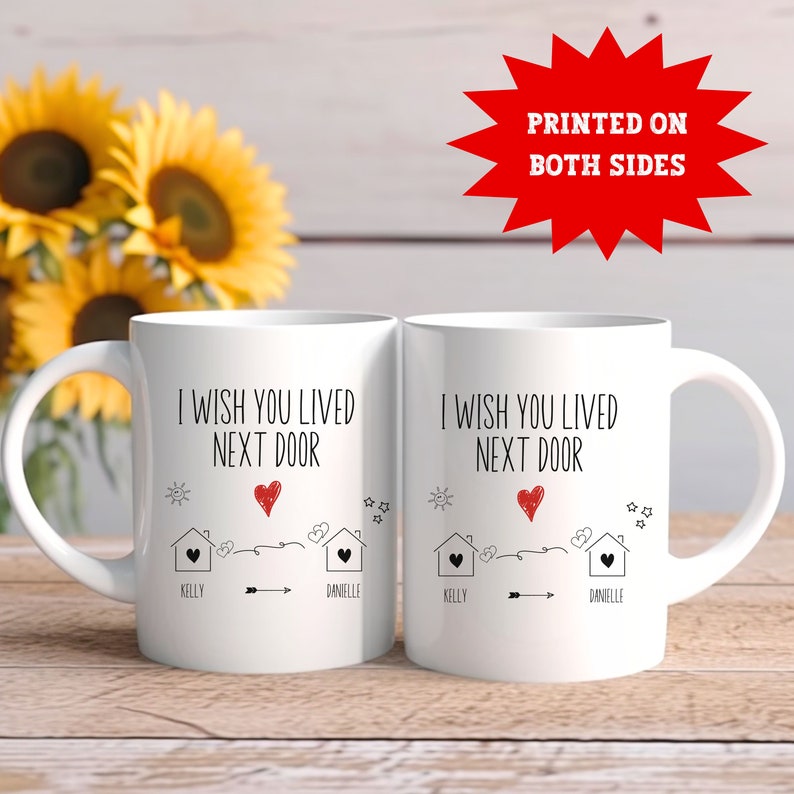 May include: Two white coffee mugs with black text that reads "I wish you lived next door" with a red heart and a drawing of two houses with the names Kelly and Danielle. The mugs are printed on both sides.