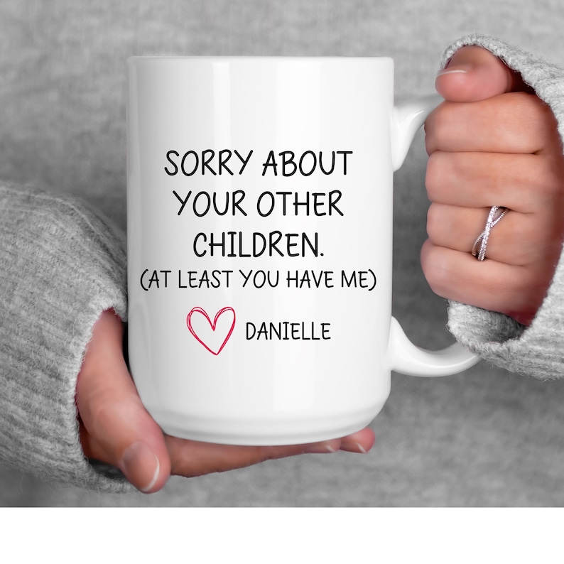 May include: White ceramic mug with black text that reads "Sorry about your other children. (At least you have me)" and a pink heart with the name "Danielle" below it.