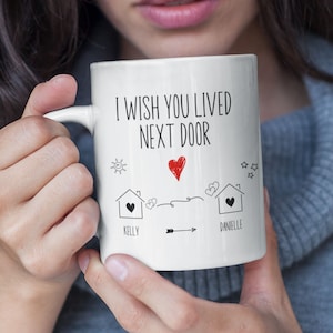 May include: White ceramic coffee mug with black text that reads "I wish you lived next door". The mug has a drawing of two houses with hearts and the names "Kelly" and "Danielle" written below.