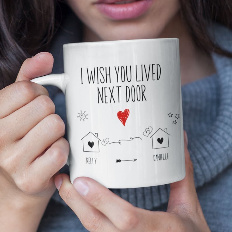 May include: White ceramic coffee mug with black text that reads "I wish you lived next door". The mug has a drawing of two houses with hearts and the names "Kelly" and "Danielle" written below.