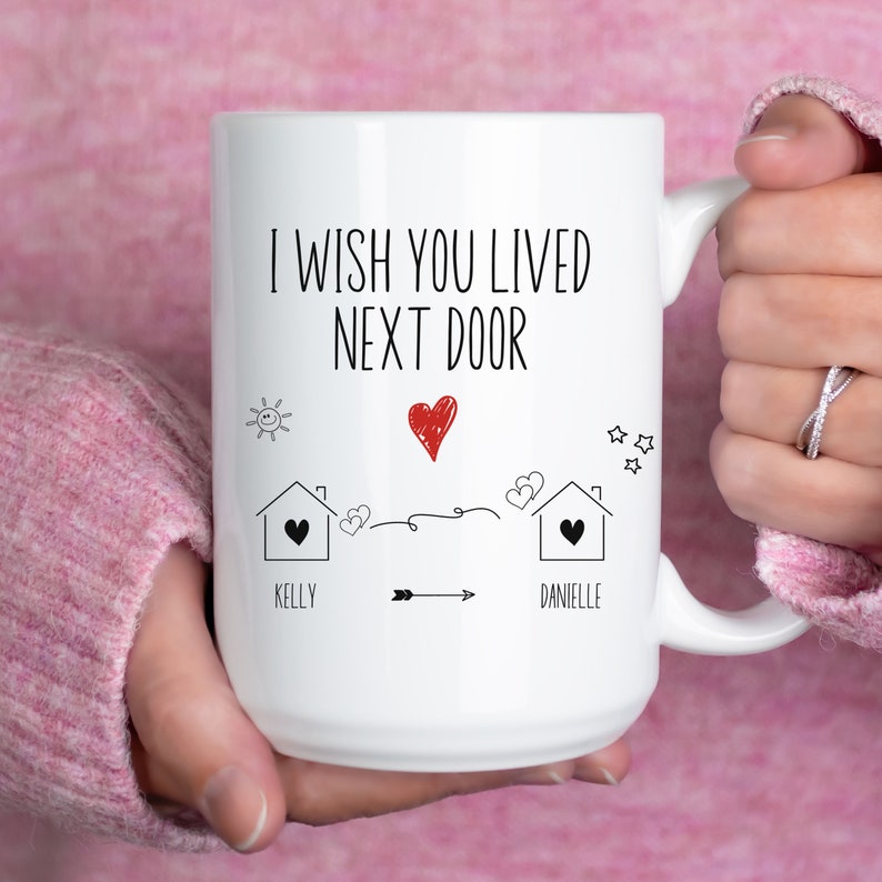 May include: White ceramic mug with black text that reads "I wish you lived next door" with a red heart and two houses with names "Kelly" and "Danielle" underneath.