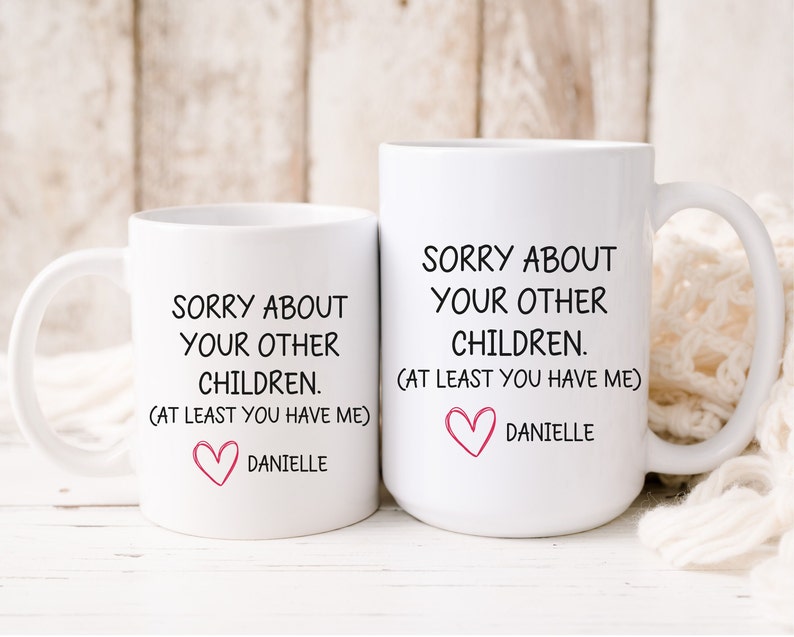 May include: Two white coffee mugs with black text that reads "Sorry about your other children. (At least you have me)" and a pink heart with the name "Danielle" below.