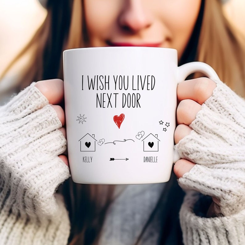 May include: White ceramic mug with a black and red design. The mug says "I wish you lived next door" with a drawing of two houses, a heart, and an arrow. The names "Kelly" and "Danielle" are written below the houses.