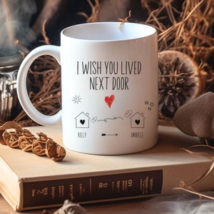 May include: White ceramic coffee mug with a black and red design. The mug says "I wish you lived next door" with two houses and hearts. The names Kelly and Danielle are written below the houses.