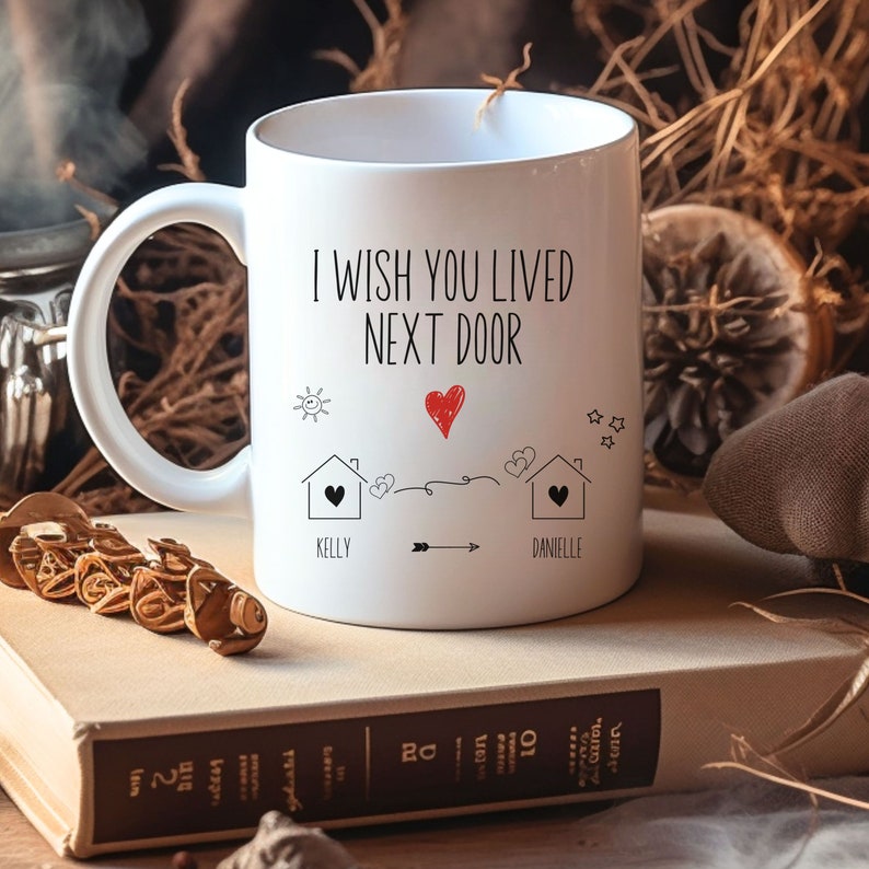 May include: White ceramic coffee mug with a black and red design. The mug says "I wish you lived next door" with two houses and hearts. The names Kelly and Danielle are written below the houses.