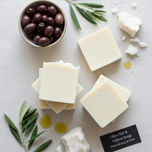 Peut inclure: Image d'un savon à l'huile d'olive et au suif, des olives et des branches d'olivier. Plusieurs savons carrés blanc cassé sont empilés et dispersés. Un petit bol d'olives noires et une branche d'olivier sont également présents. Une étiquette noire indique "Olive Oil & Tallow Soap - Nourishing & Pure."