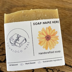 Puede incluir: Una pastilla de jabón artesanal con una etiqueta blanca con una ilustración de girasol y el texto "SOAP NAME HERE". La etiqueta también incluye el texto "All Natural", "Plant Based" y "Sustainable Ingredients".
