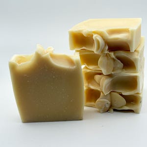 May include: A stack of four rectangular bars of cream-coloured soap with a smooth, solid texture. One additional bar of the same cream-coloured soap stands upright to the left of the stack, displaying a slightly textured top edge and a solid body.