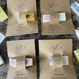 May include: Four sample bars of handmade soap are presented. Each is attached to a brown card with a heart and the text "Soap Name Handmade Soap". The soap bars are various colours, including yellow, pink, and white. The cards also say "Enjoy this sample bar of soap."