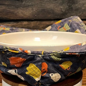 Puede incluir: Un bol de cerámica blanca está sentado dentro de un calientaplatos de tela azul con un patrón colorido de tazas y teteras. El calientaplatos está hecho de tela de algodón y tiene un diseño acolchado.