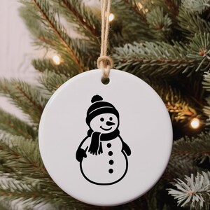 May include: A white, round Christmas ornament featuring a black snowman design. The snowman wears a hat, scarf, and mittens. The ornament hangs from a natural twine string, set against a backdrop of a Christmas tree.