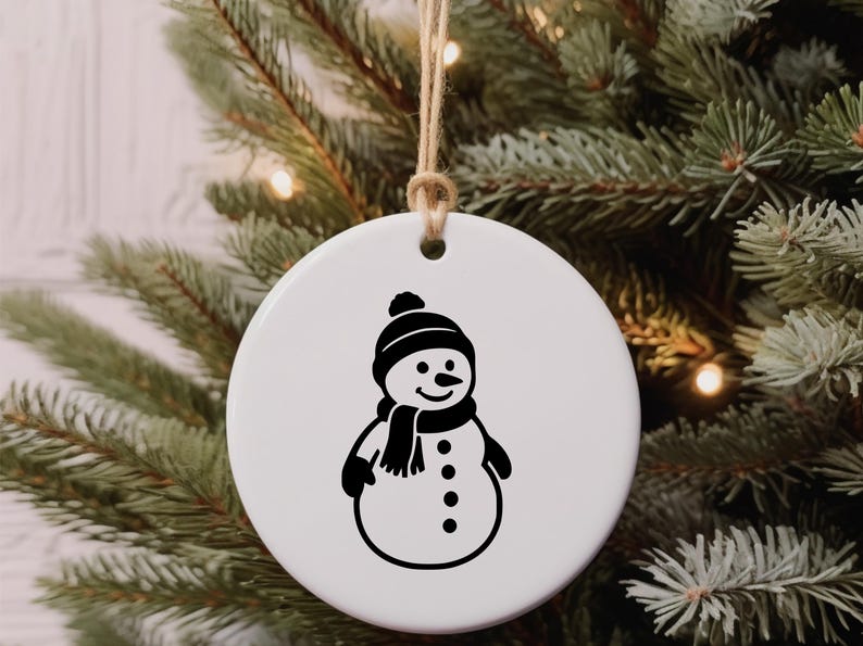 May include: A white, round Christmas ornament featuring a black snowman design. The snowman wears a hat, scarf, and mittens. The ornament hangs from a natural twine string, set against a backdrop of a Christmas tree.