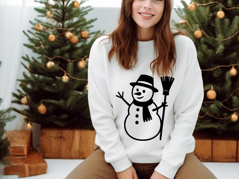 May include: White sweatshirt featuring a black and white snowman graphic. The snowman wears a hat, scarf, and holds a broom. The design is centered on the front of the long-sleeved top. The background includes Christmas trees with gold ornaments.