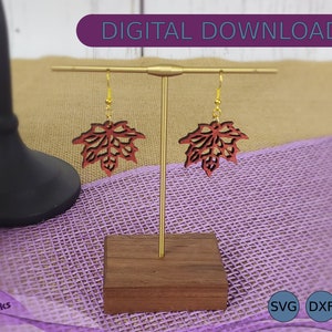 May include: A pair of red maple leaf earrings on a gold stand. The earrings are made of wood and have a delicate, intricate design. The stand is made of wood and has a rectangular base. The earrings are on a purple fabric background. SVG, DXF, and EPS files are available for download.