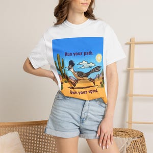 Peut inclure: Un t-shirt blanc à manches courtes avec un motif représentant un géocoucou portant des bottes de cow-boy courant dans un paysage désertique avec des cactus, un soleil et des nuages. Le texte indique Run your path. Own your speed.