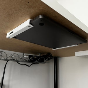May include: A black laptop computer is mounted under a white shelf with a white bracket. The laptop is facing down with the screen closed. The shelf is attached to a brown surface.