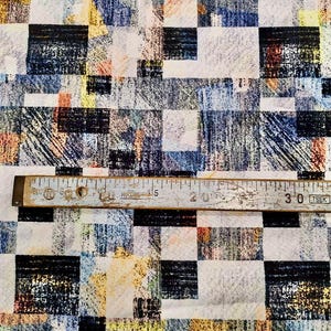 以下が含まれることがあります： 黒、白、青、黄色、オレンジ色の正方形の幾何学模様の生地。センチメートル単位の測定値が記載された木製の定規が生地の上に置かれています。