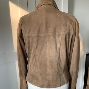 K&ouml;nnte beinhalten: Eine braune Wildlederjacke mit Kragen und langen &Auml;rmeln. Die Jacke hat eine figurbetonte Silhouette und eine verk&uuml;rzte L&auml;nge.