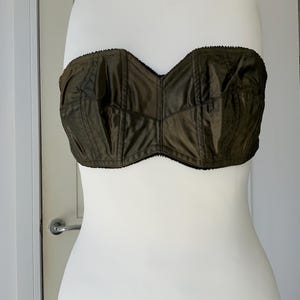 May include: A strapless bra made of dark green satin with black trim. The bra has a boned structure and a sweetheart neckline.