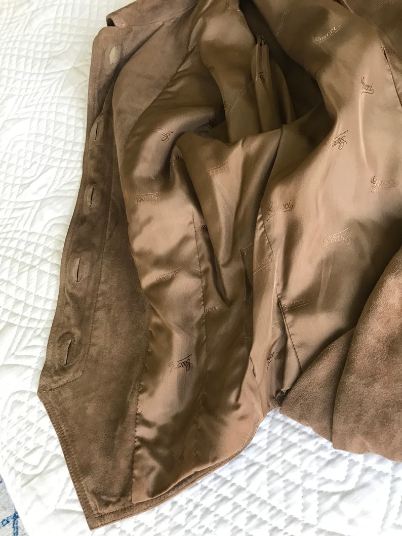 K&ouml;nnte beinhalten: Eine braune Wildlederjacke mit braunem Satin-Futter. Das Futter ist mit dem Wort "Gucci" durchgehend bedruckt.