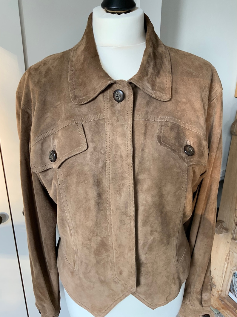 K&ouml;nnte beinhalten: Eine braune Wildlederjacke mit Knopfleiste und zwei Pattentaschen. Die Jacke hat einen spitzen Saum und lange &Auml;rmel.