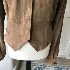 K&ouml;nnte beinhalten: Eine braune Wildlederjacke mit spitzem Saum und langen &Auml;rmeln. Die Jacke hat einen Knopfverschluss und einen Kragen.