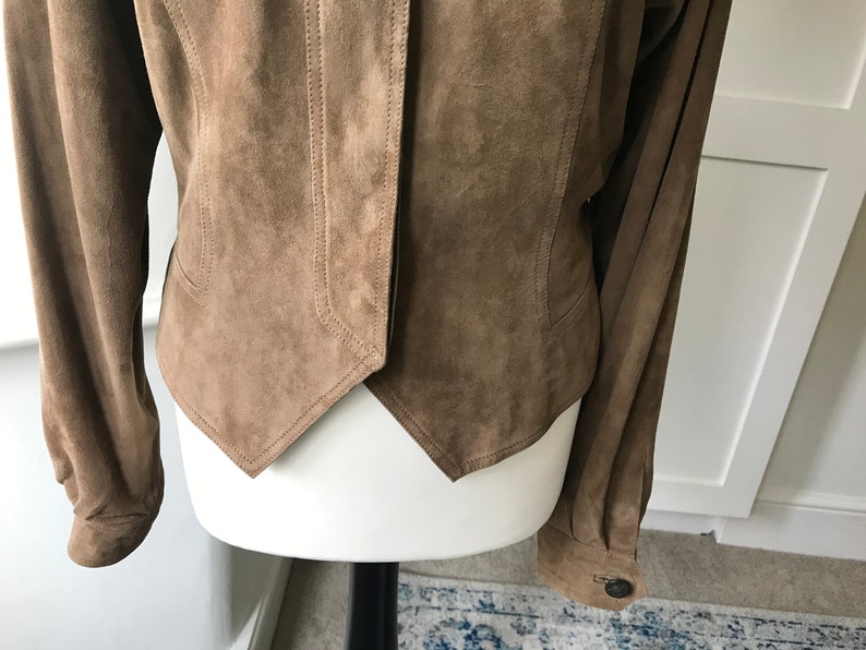 K&ouml;nnte beinhalten: Eine braune Wildlederjacke mit spitzem Saum und langen &Auml;rmeln. Die Jacke hat einen Knopfverschluss und einen Kragen.