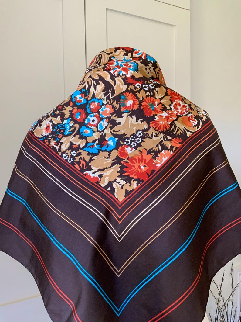 K&ouml;nnte beinhalten: Ein quadratischer Schal mit braunem Hintergrund und einem Blumenmuster in Rot, Blau und Beige. Der Schal hat einen gestreiften Rand mit roten, wei&szlig;en und blauen Linien. Der Schal ist aus seidigem Material.