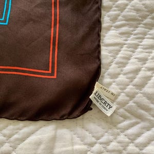 K&ouml;nnte beinhalten: Brauner Seidenschal mit geometrischem Design in Orange und Blau. Der Schal hat ein Etikett mit der Aufschrift "LIBERTY MADE IN ITALY". Der Schal ist aus einem weichen, flie&szlig;enden Material gefertigt.