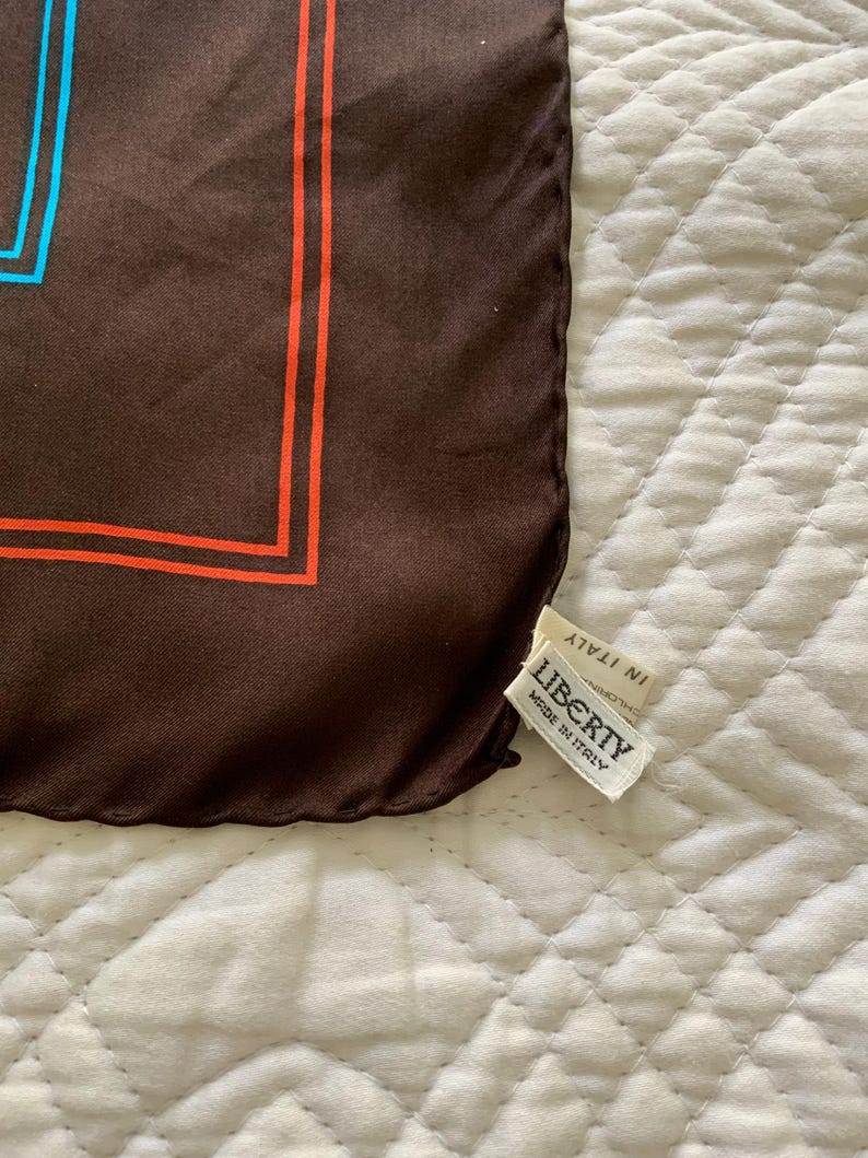 K&ouml;nnte beinhalten: Brauner Seidenschal mit geometrischem Design in Orange und Blau. Der Schal hat ein Etikett mit der Aufschrift "LIBERTY MADE IN ITALY". Der Schal ist aus einem weichen, flie&szlig;enden Material gefertigt.