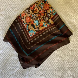 K&ouml;nnte beinhalten: Ein quadratisches, braunes Seidentuch mit einem Blumenmuster in Orange, Beige und Blau. Der Schal hat einen gestreiften Rand mit blauen und roten Akzenten. Der Schal ist gefaltet und liegt auf einer wei&szlig;en Oberfl&auml;che.