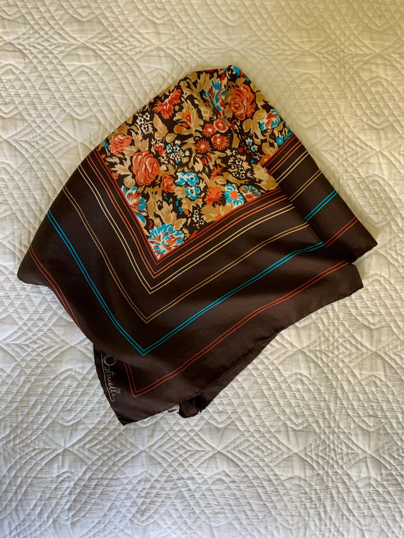 K&ouml;nnte beinhalten: Ein quadratisches, braunes Seidentuch mit einem Blumenmuster in Orange, Beige und Blau. Der Schal hat einen gestreiften Rand mit blauen und roten Akzenten. Der Schal ist gefaltet und liegt auf einer wei&szlig;en Oberfl&auml;che.