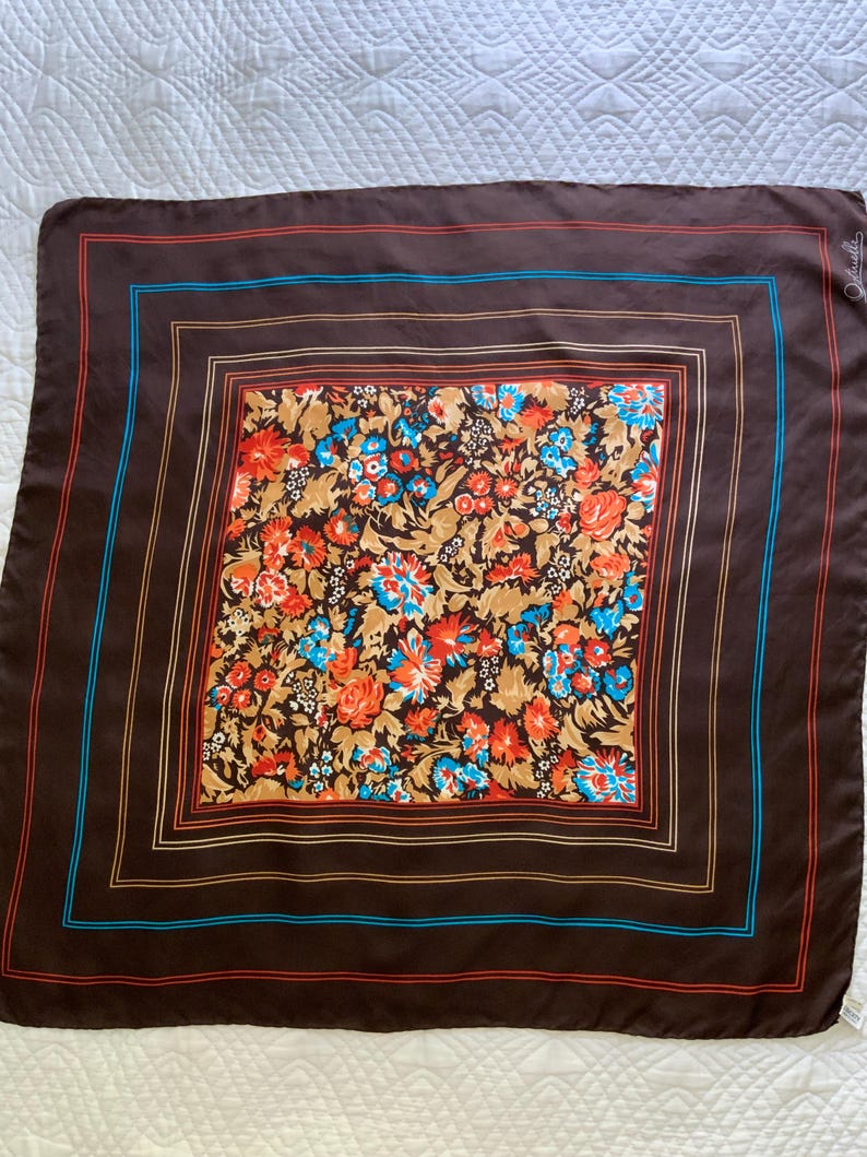 K&ouml;nnte beinhalten: Ein quadratisches braunes Seidentuch mit floralem Muster. Die Mitte zeigt eine lebendige Mischung aus orangefarbenen, blauen und beigen Blumen. Der Schal hat einen Rand mit roten und blauen Streifen. Der Schal ist wahrscheinlich Vintage.
